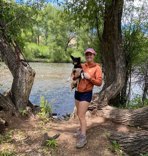 Eva stands in front of a river holding her dog. There are cottonwood trees on either side of her.